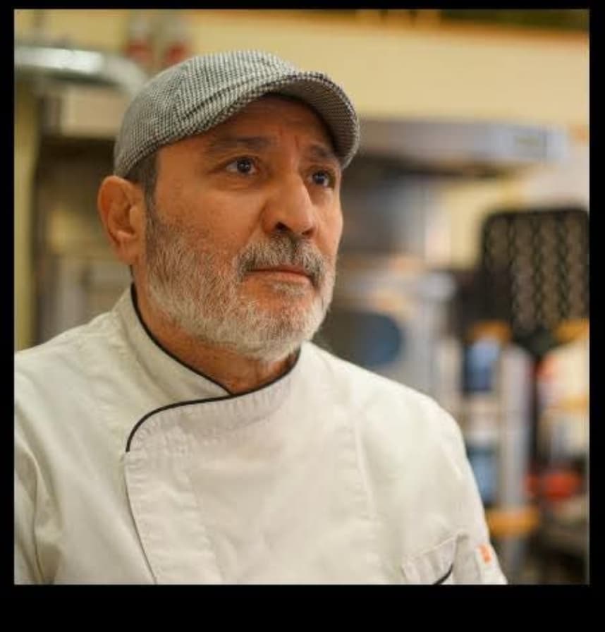 Cocinero con barba canosa y gorra gris mirando fijamente con su chaqueta blanca de chef.