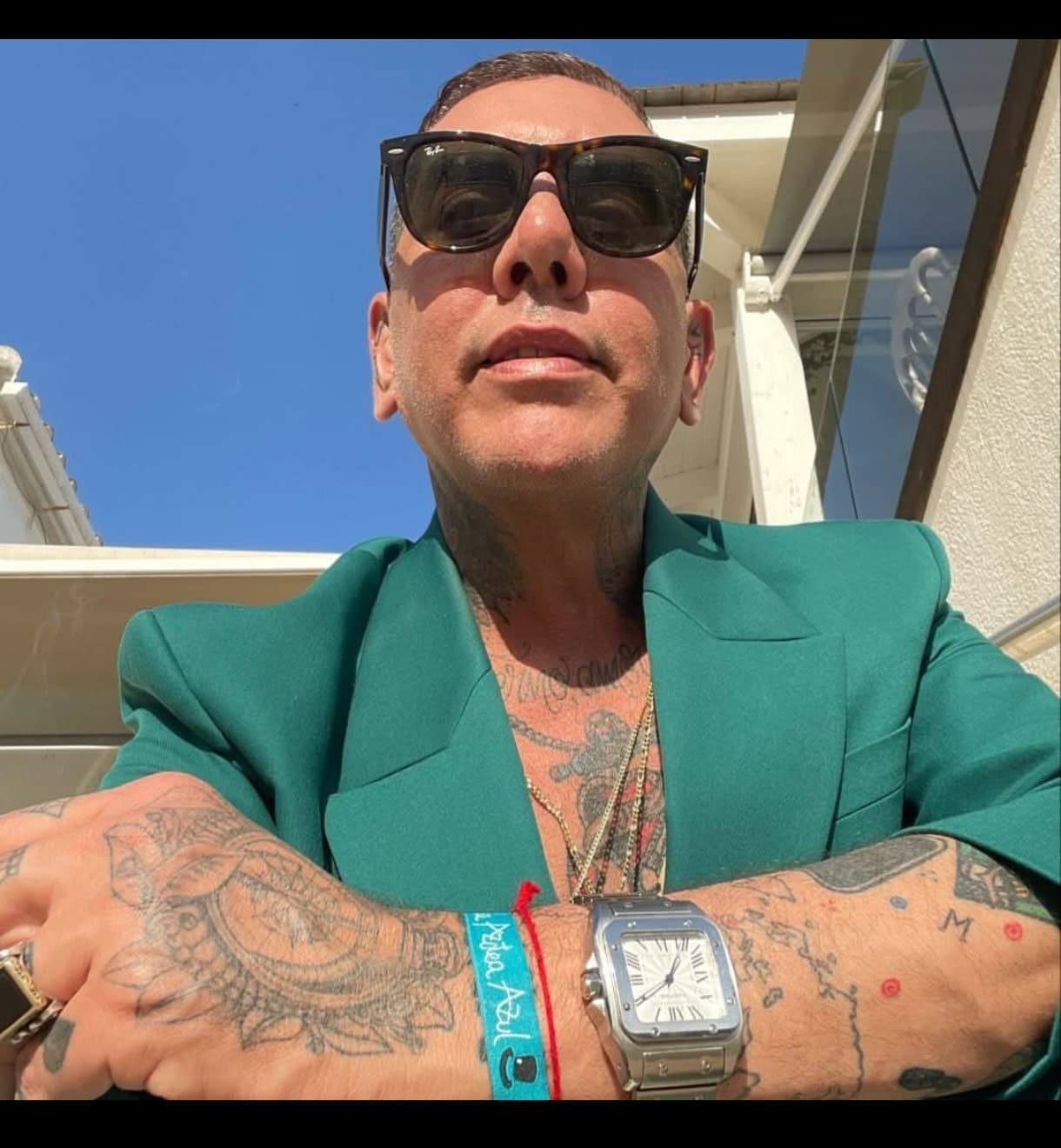Hombre tatuado con gafas de sol y chaqueta verde posando bajo un cielo azul.